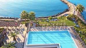 A swimming pool with sun loungers and palm trees, overlooking a rocky coastline. - Crowne Plaza Limassol by IHG (Limassol)