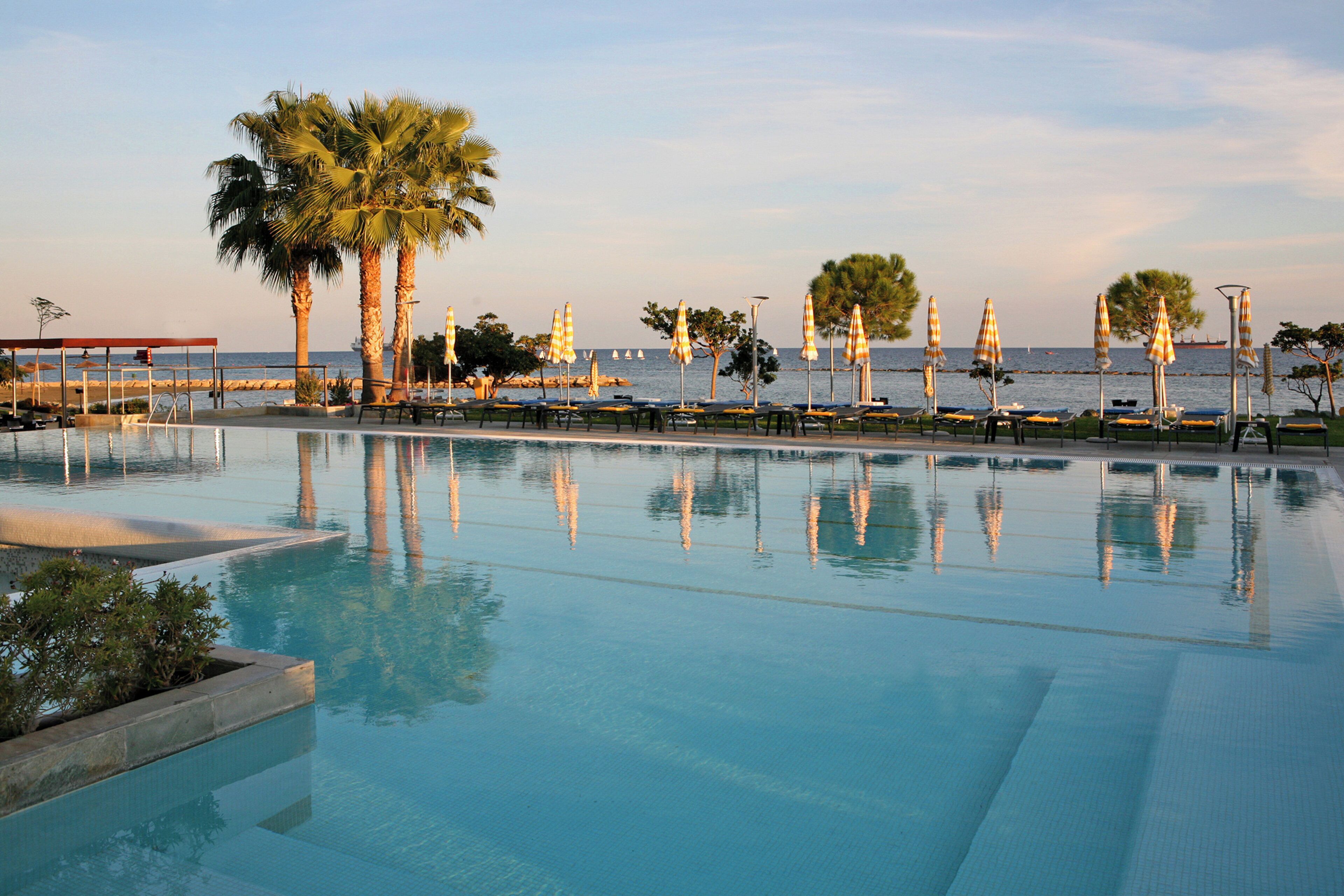 A pool area with lounge chairs and umbrellas, palm trees, and a view of the sea.