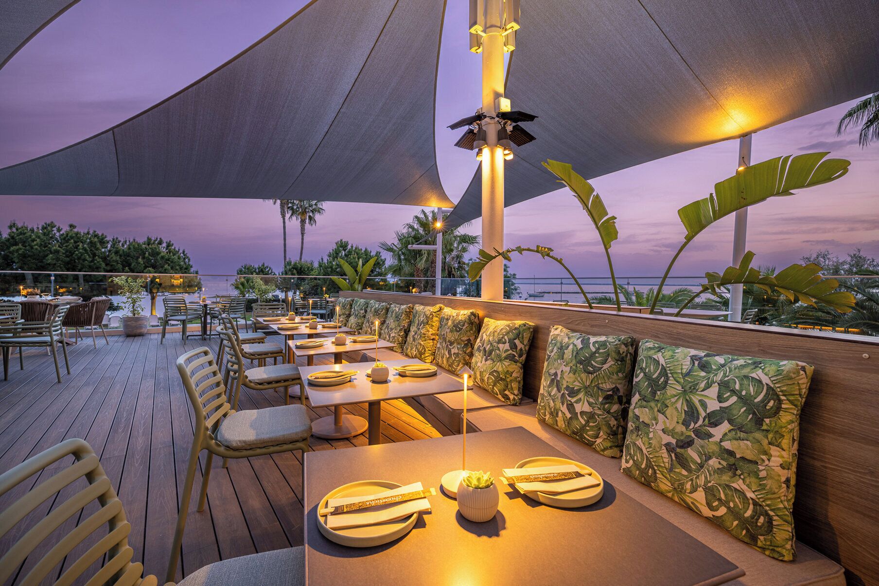 An outdoor dining area with wooden tables and chairs, a canopy, and a view of the sea.