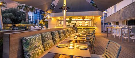 Un espace extérieur aménagé pour les repas, avec des tables et des bancs en bois, un coin bar doté d’un comptoir, et un grand parasol offrant de l’ombre.