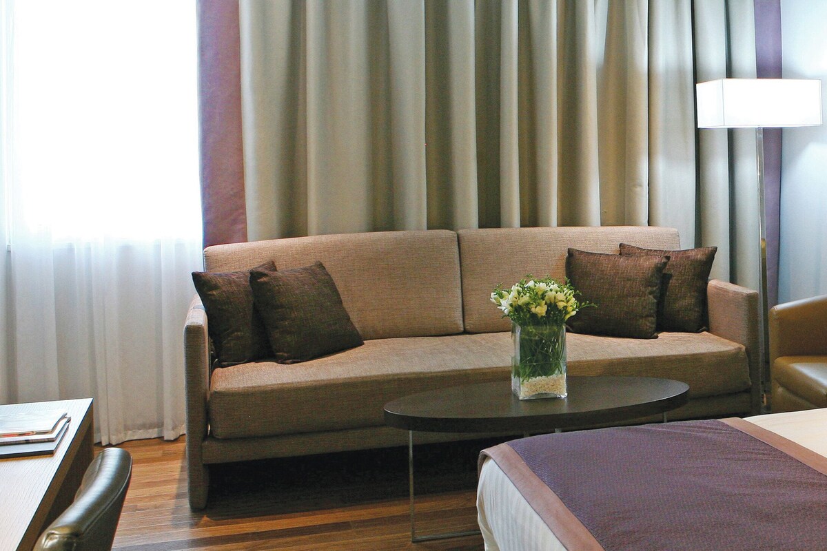 Une chambre d’hôtel avec un canapé, une petite table ronde ornée d’un vase de fleurs, une lampe et un lit.