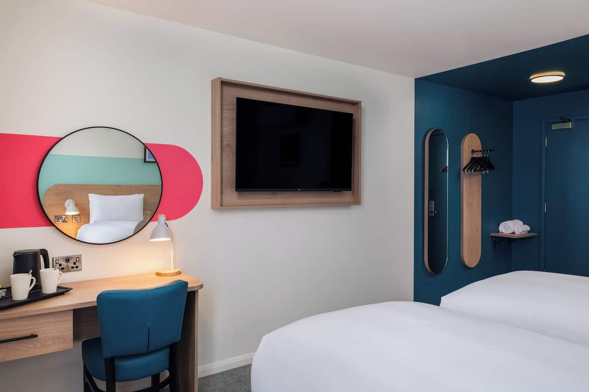 A hotel room with a bed, a desk with a chair, a TV, a mirror, and a shower area.