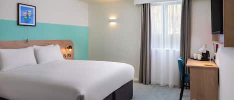 A hotel room with a large bed, a desk with a lamp, a chair, and a television on the wall.