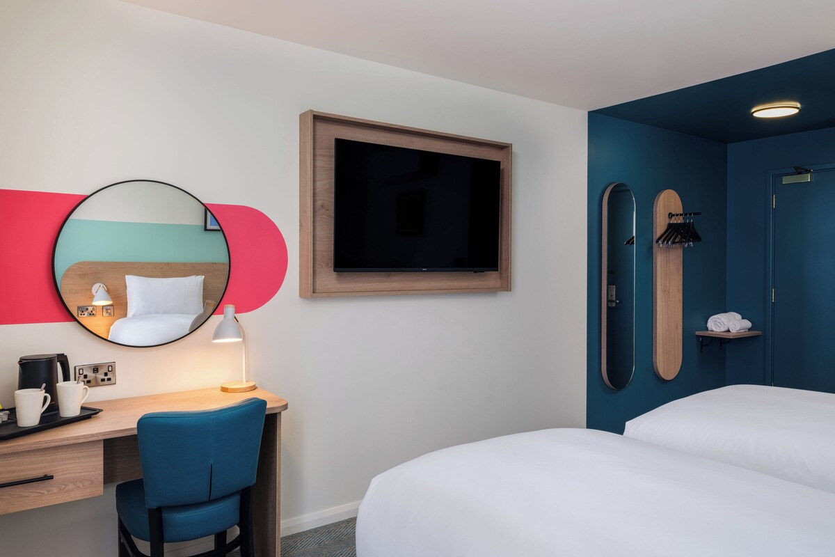 Habitación de hotel con cama, escritorio con silla, televisor, espejo y zona de ducha.