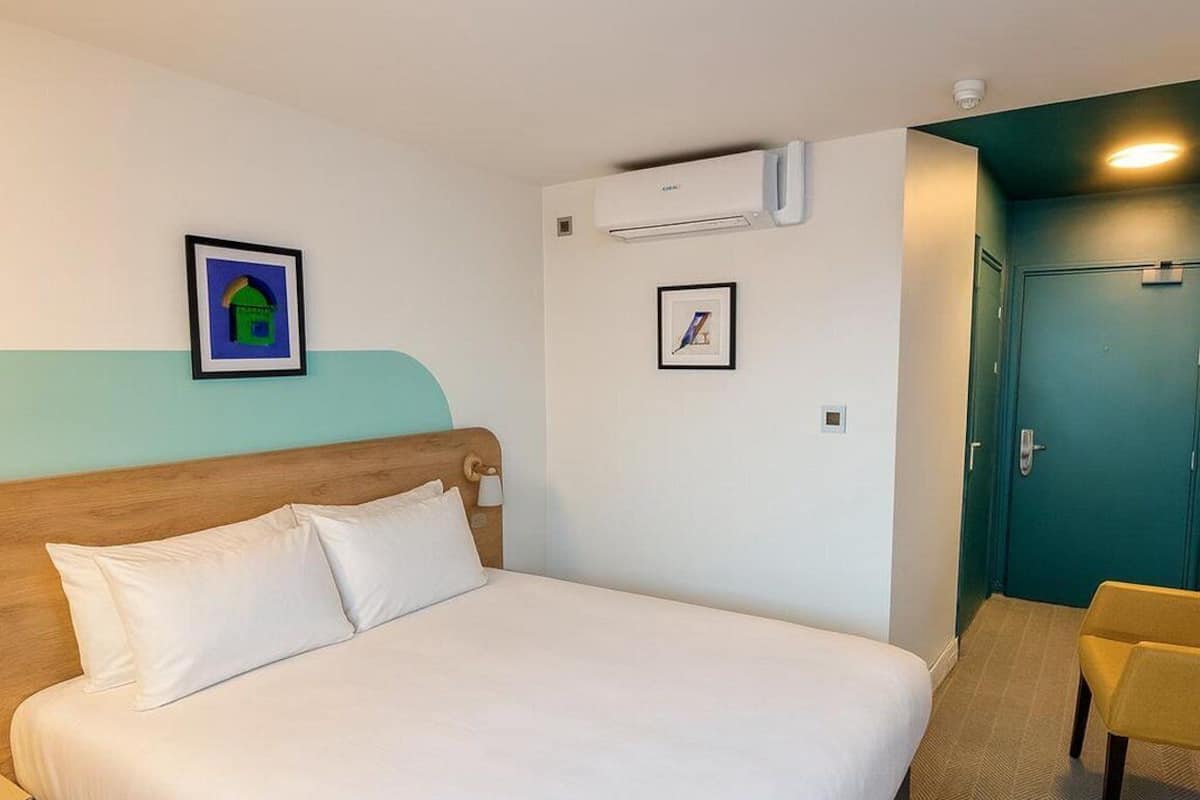 A hotel room with a bed, a chair, an air conditioning unit, and two framed pictures on the wall.