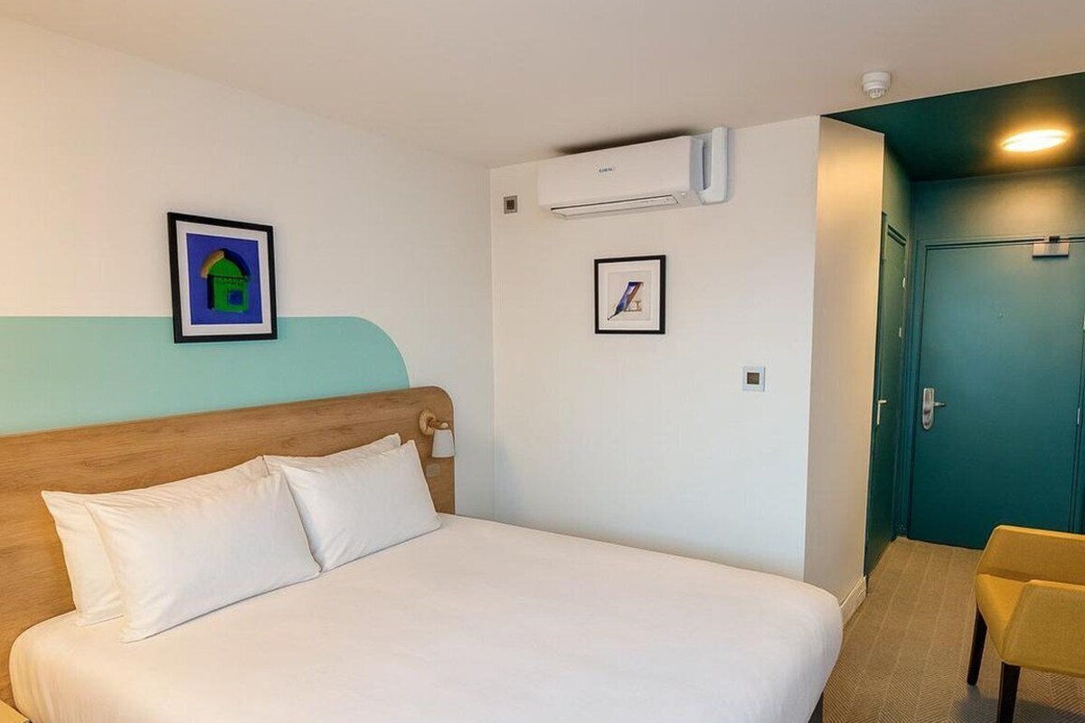 Habitación de hotel con una cama, una silla, un equipo de aire acondicionado y dos cuadros en la pared.