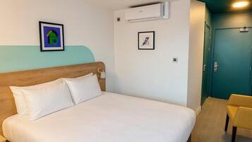 A hotel room with a bed, a chair, an air conditioning unit, and two framed pictures on the wall.