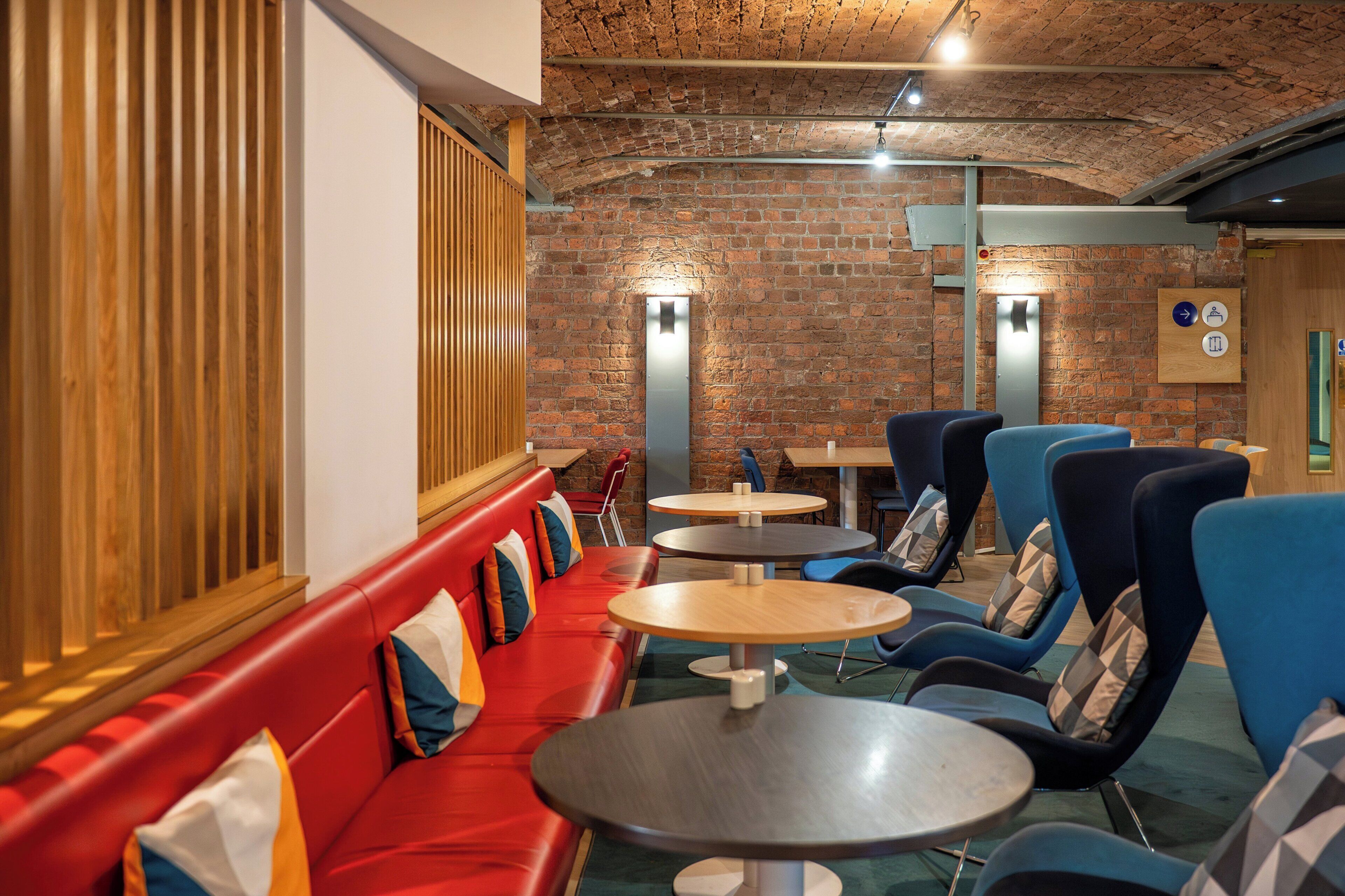 A modern interior with a brick wall, wooden slats, red seating, and round tables.