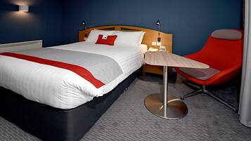 A hotel room with a large bed, a red chair, a small table, and a lamp.