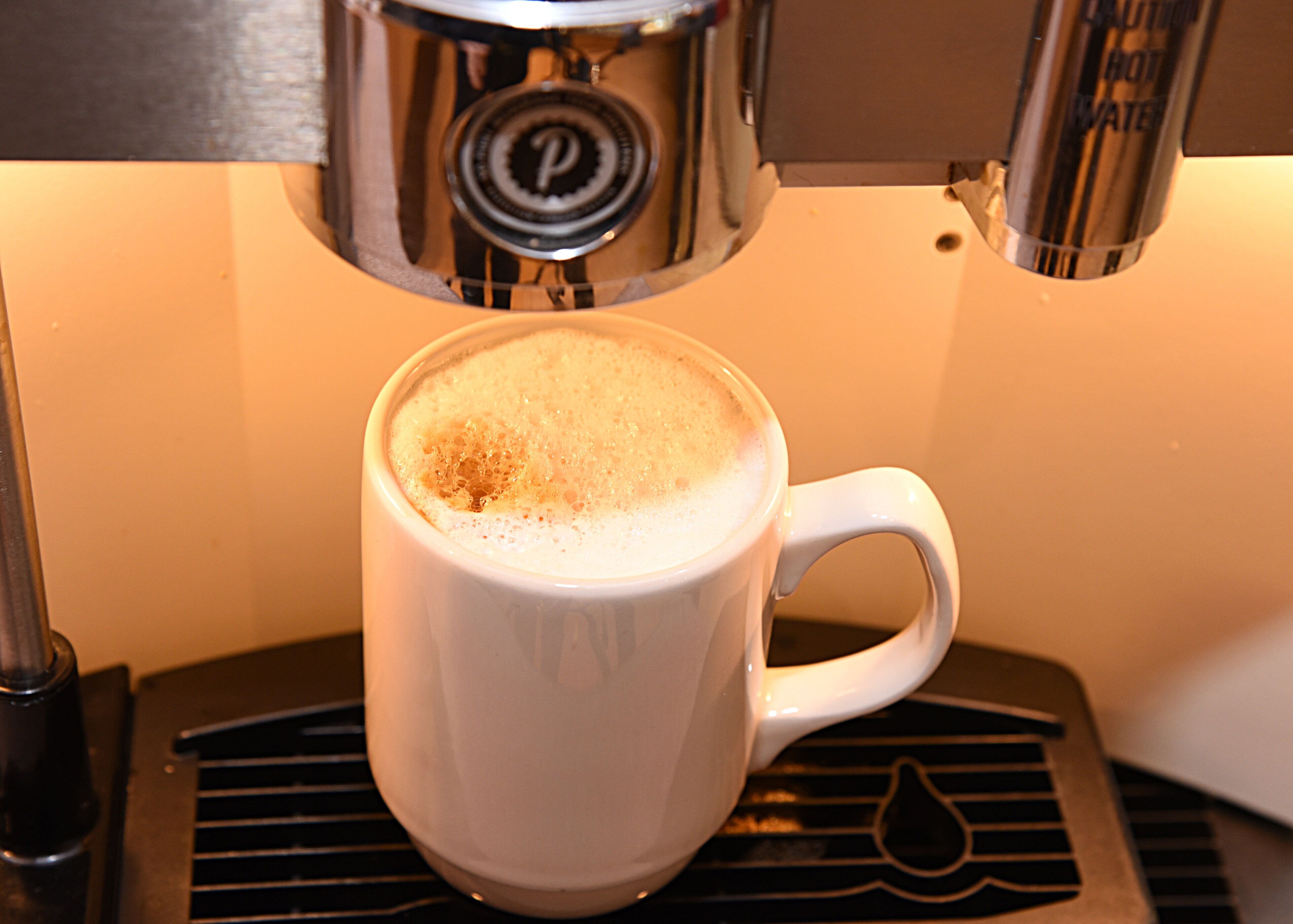 A white cup with a frothy top, placed on a black surface with a logo.