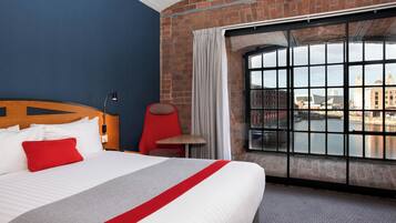 Ett hotellrum med en stor säng, en röd stol, ett litet bord och ett fönster med utsikt över en flod och byggnader.
