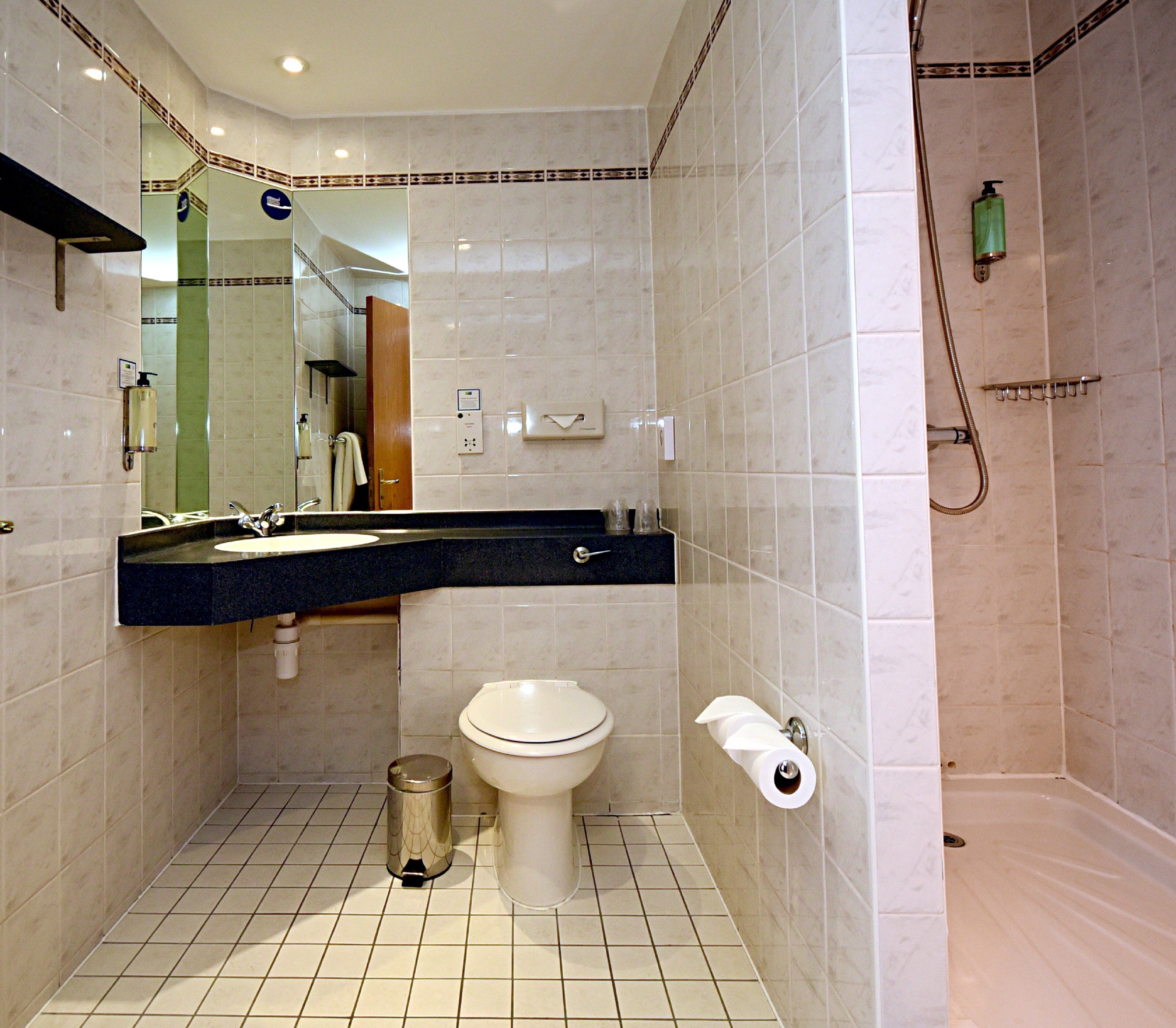 A bathroom with a toilet, sink, mirror, shower, and a trash can.