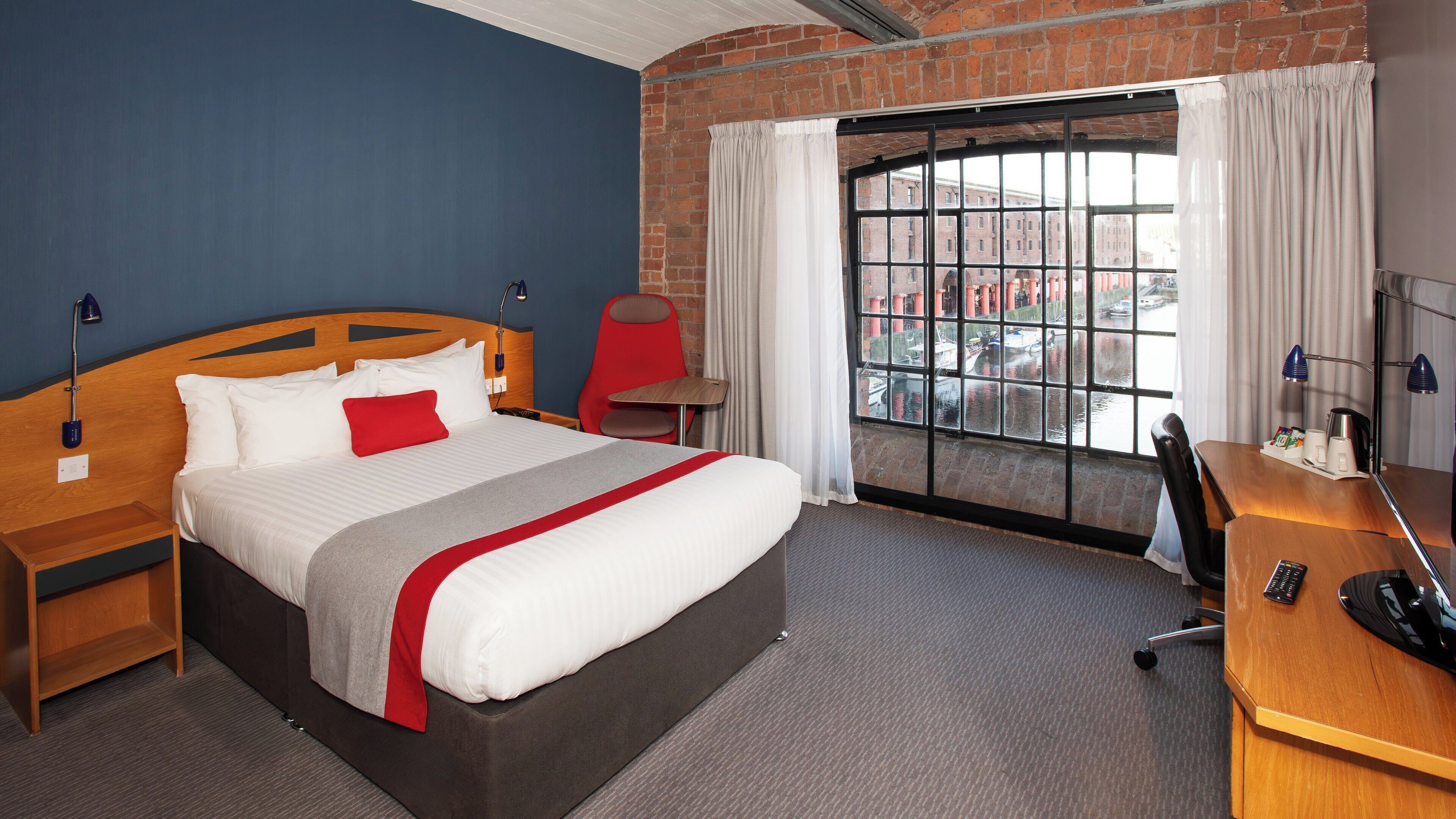 Habitación de hotel con una cama grande, un escritorio, una silla, un ventanal con vistas a edificios y una pared de ladrillo.