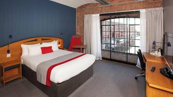 Habitación de hotel con una cama grande, un escritorio, una silla, un ventanal con vistas a edificios y una pared de ladrillo.