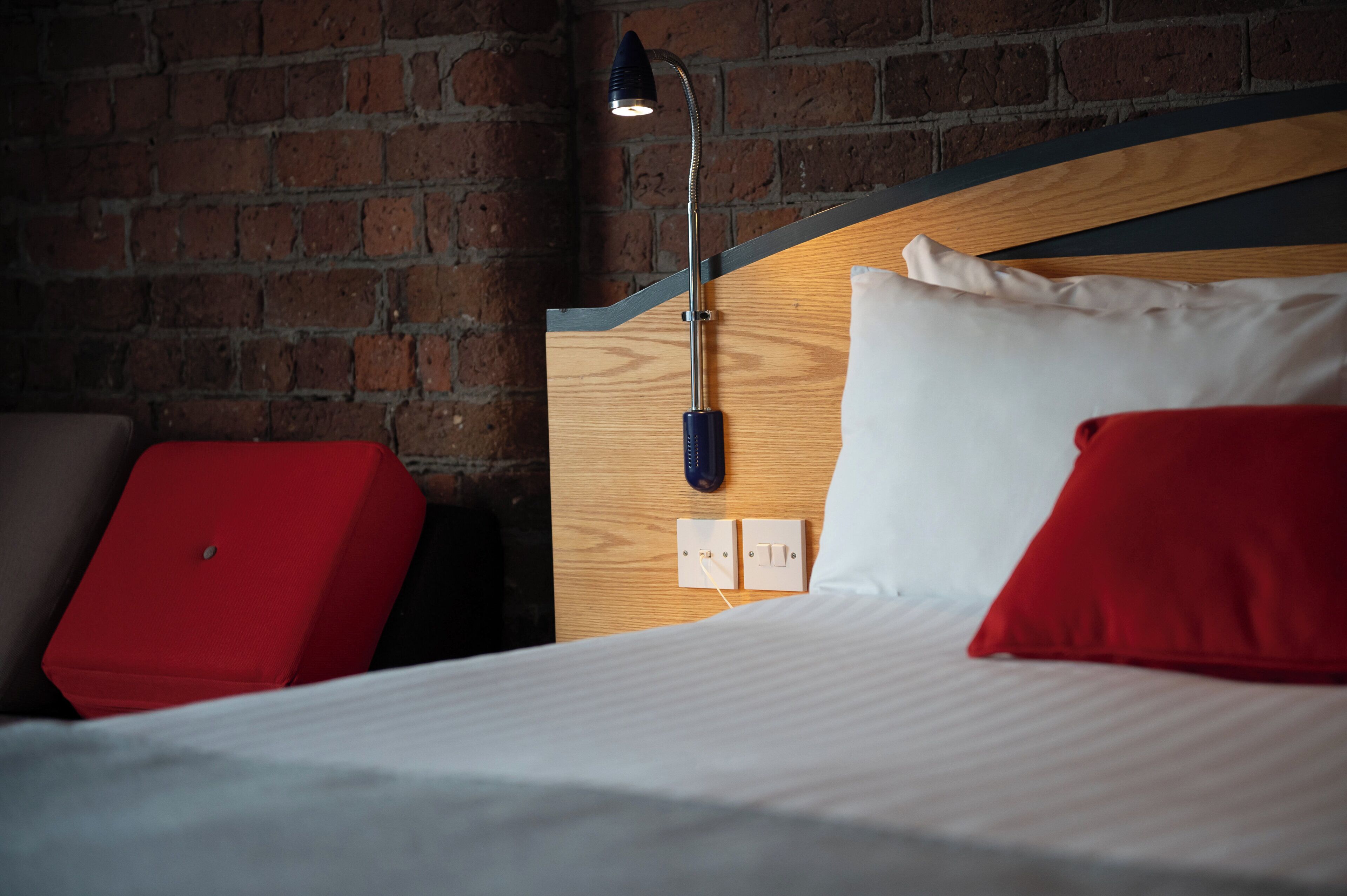 A hotel room with a brick wall, a wooden headboard, a bed with white bedding, red pillows, and a bedside lamp.