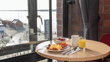 A breakfast table with a plate of food, a glass of orange juice, and a cup, set against a window with a view of a river and buildings.