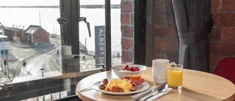 Una mesa de desayuno con un plato de comida, un vaso de jugo de naranja y una taza, frente a una ventana con vista al río y edificios.