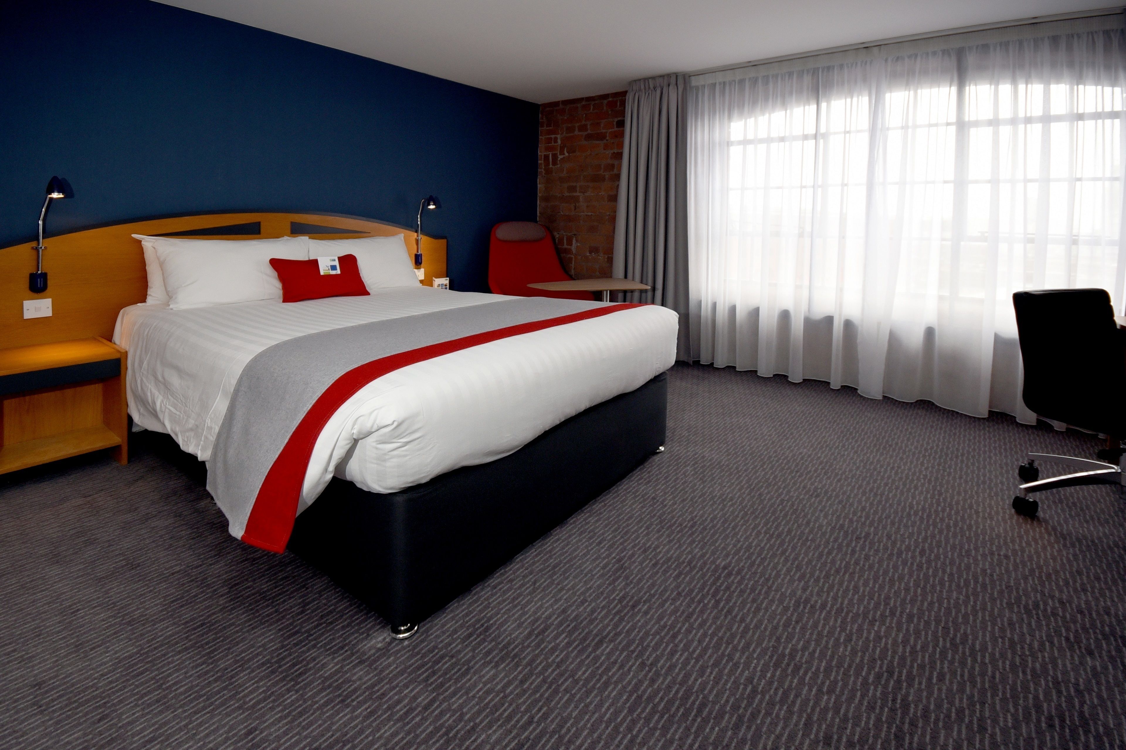 A hotel room with a large bed, a wooden headboard, a red accent pillow, a red chair, a small table, and a large window with sheer curtains.