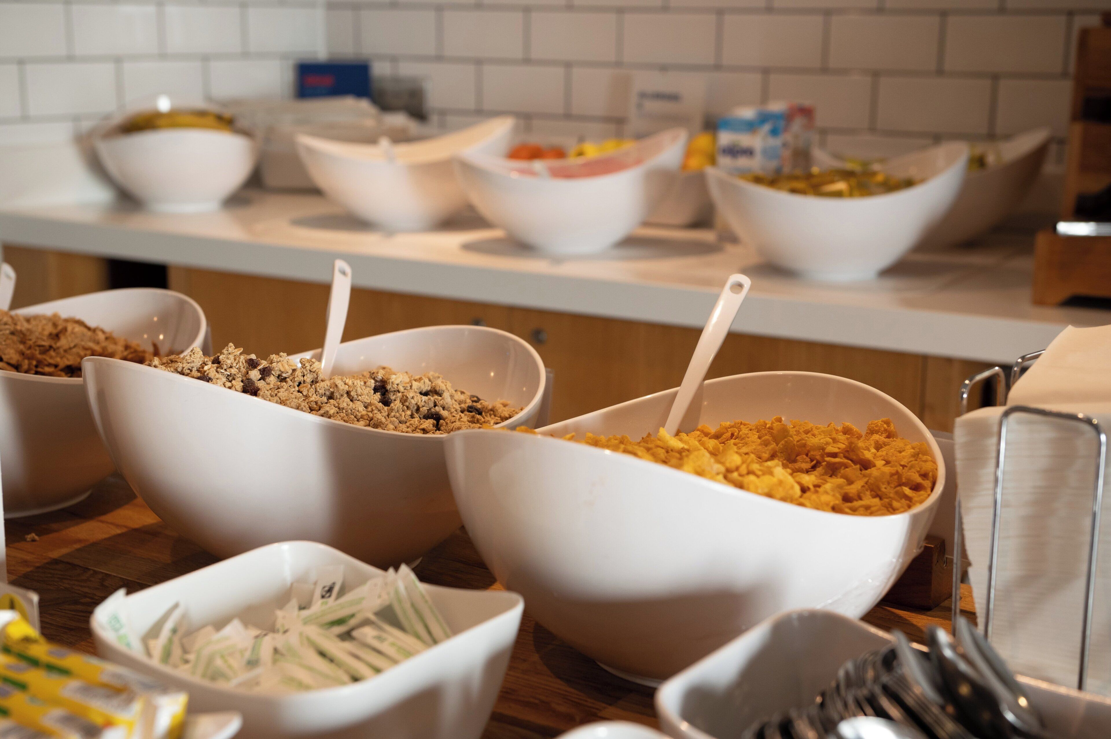 Un bufé con distintos alimentos servidos en recipientes blancos, como cereales y verduras ralladas.