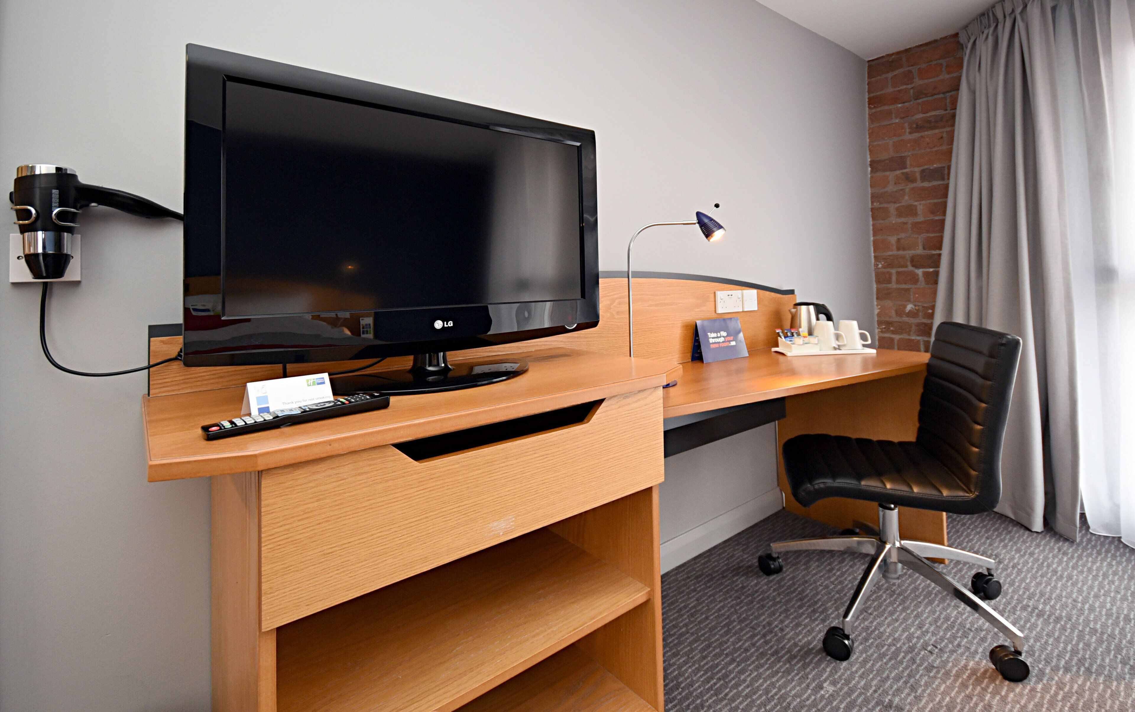 A hotel room with a wooden desk, a black office chair, a television, a remote control, and a coffee maker.