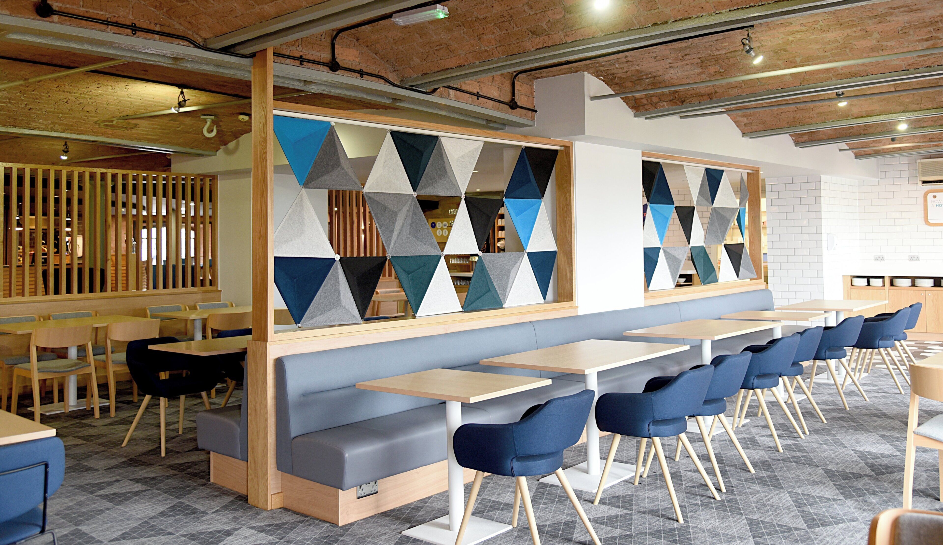 A modern restaurant interior with long communal tables, blue upholstered chairs, and geometric patterned partitions.
