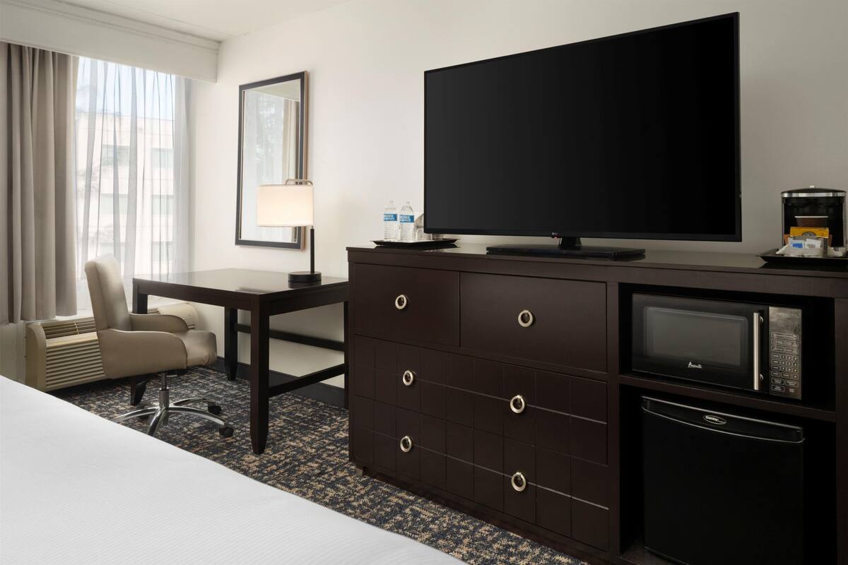 A hotel room with a bed, a desk with a chair, a dresser with a television, a microwave, and a refrigerator.