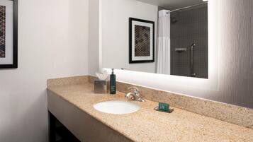 A bathroom with a granite countertop sink, a mirror, a framed artwork, and a shower area.