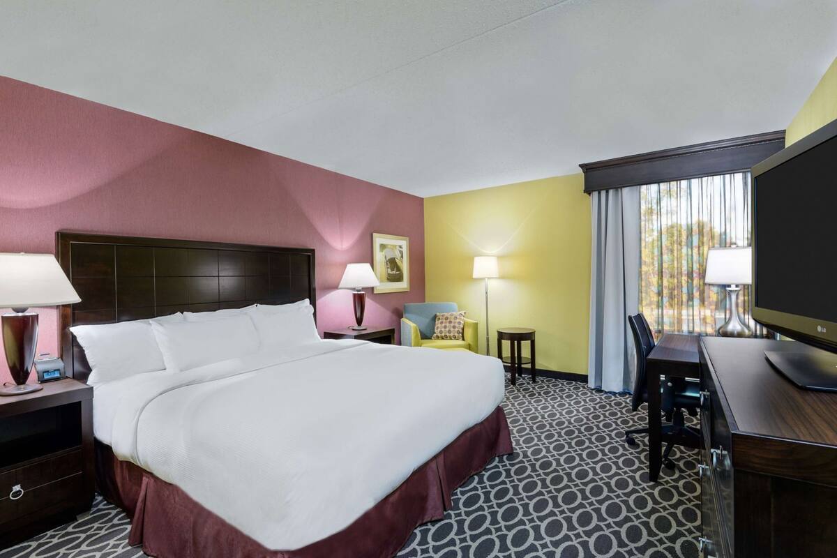 A hotel room with a large bed, a desk with a TV, a chair, a lamp, and a small table.