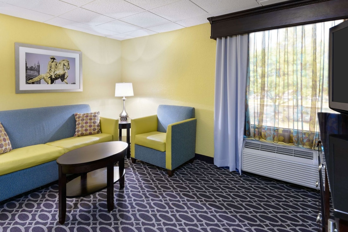A hotel room with a yellow wall, a framed picture, a sofa, a chair, a small table, a lamp, and a window with curtains.