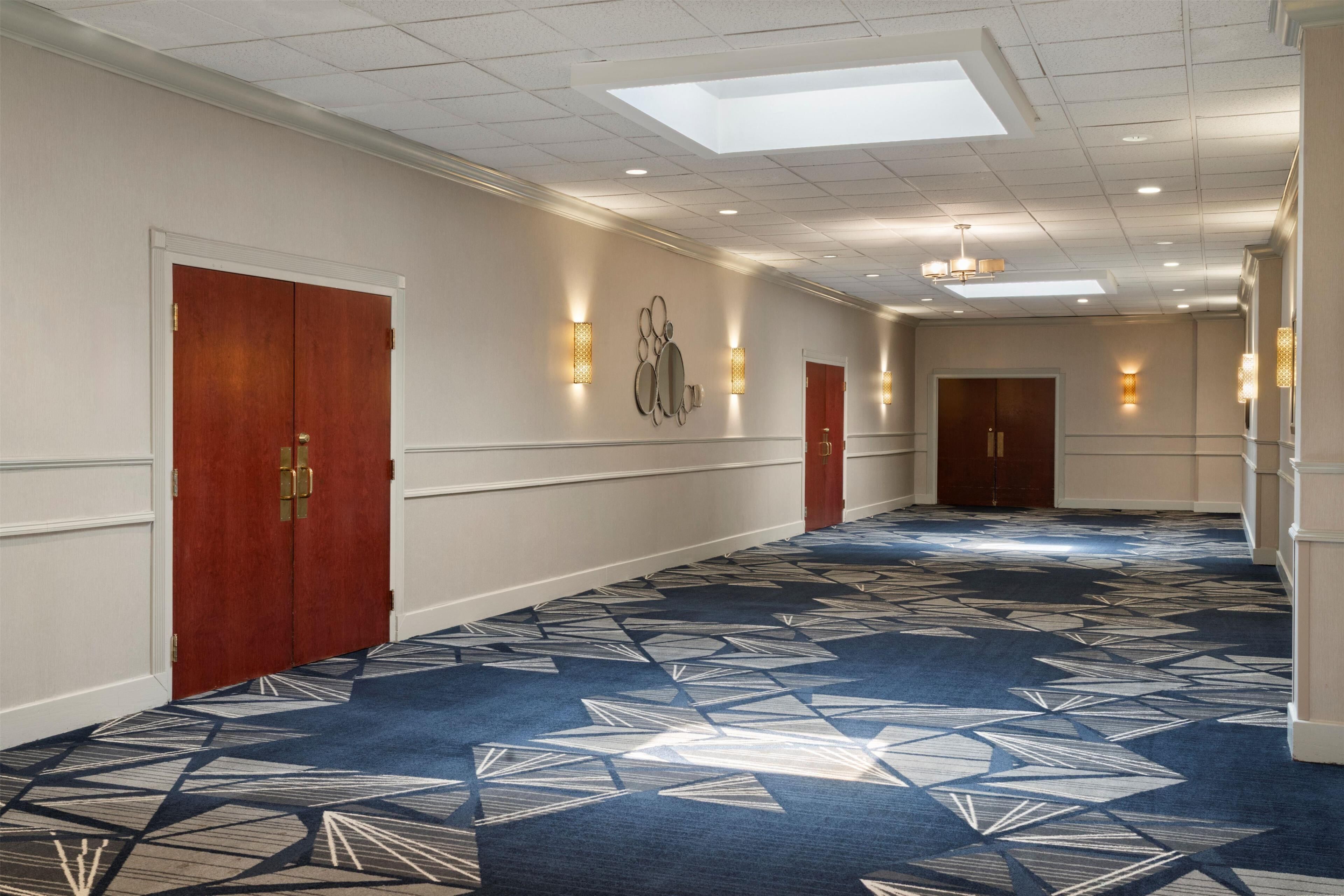 A hotel corridor with patterned carpet, wooden doors, and wall sconces.