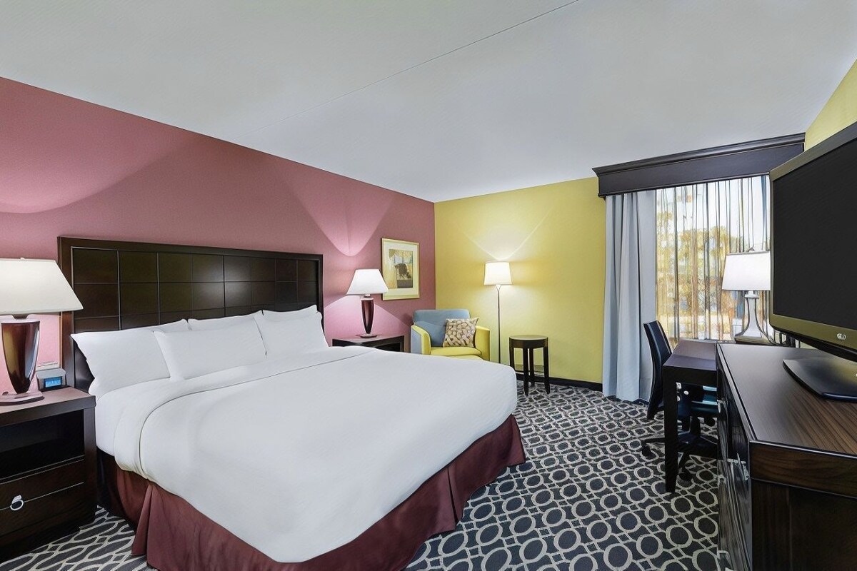 A hotel room with a large bed, bedside tables, a desk with a TV, a chair, and a window with curtains.