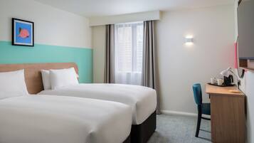 Ein Hotelzimmer mit zwei Betten, einem Schreibtisch mit Stuhl, einem Fenster mit Vorhängen und einem gerahmten Bild an der Wand.