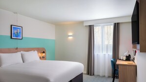 A hotel room with a large bed, a desk with a lamp, a chair, and a TV mounted on the wall.