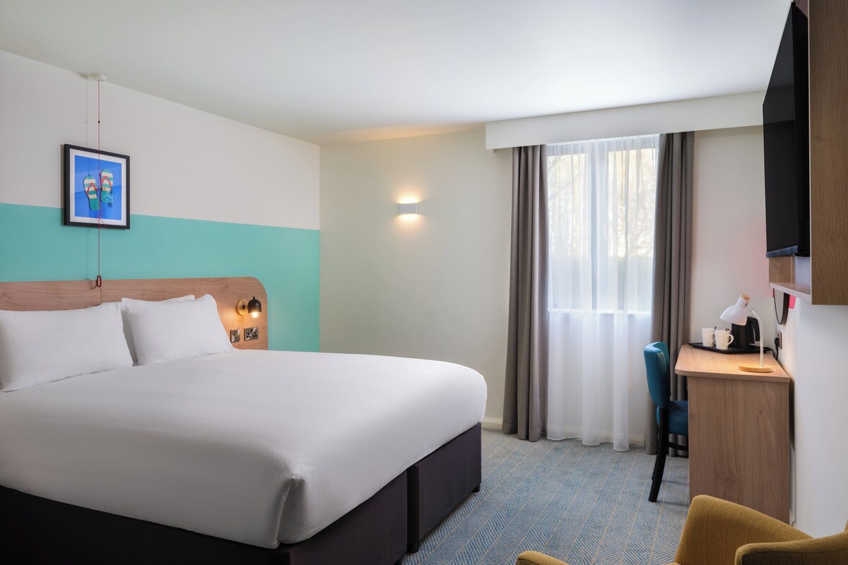 Habitación de hotel con una cama grande, un escritorio con lámpara, una silla y un televisor montado en la pared.