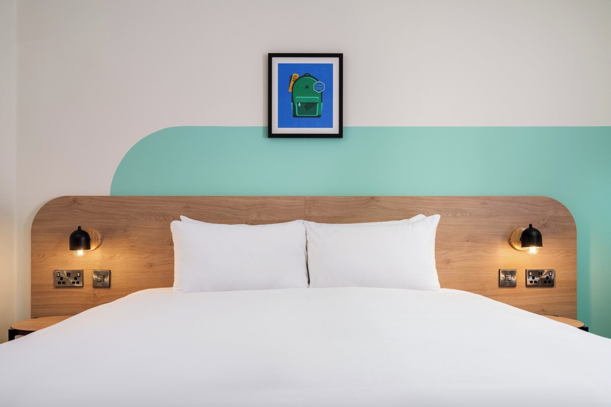 Una cama bien hecha con sábanas blancas, cabecera de madera y dos lámparas de lectura fijadas a la pared.