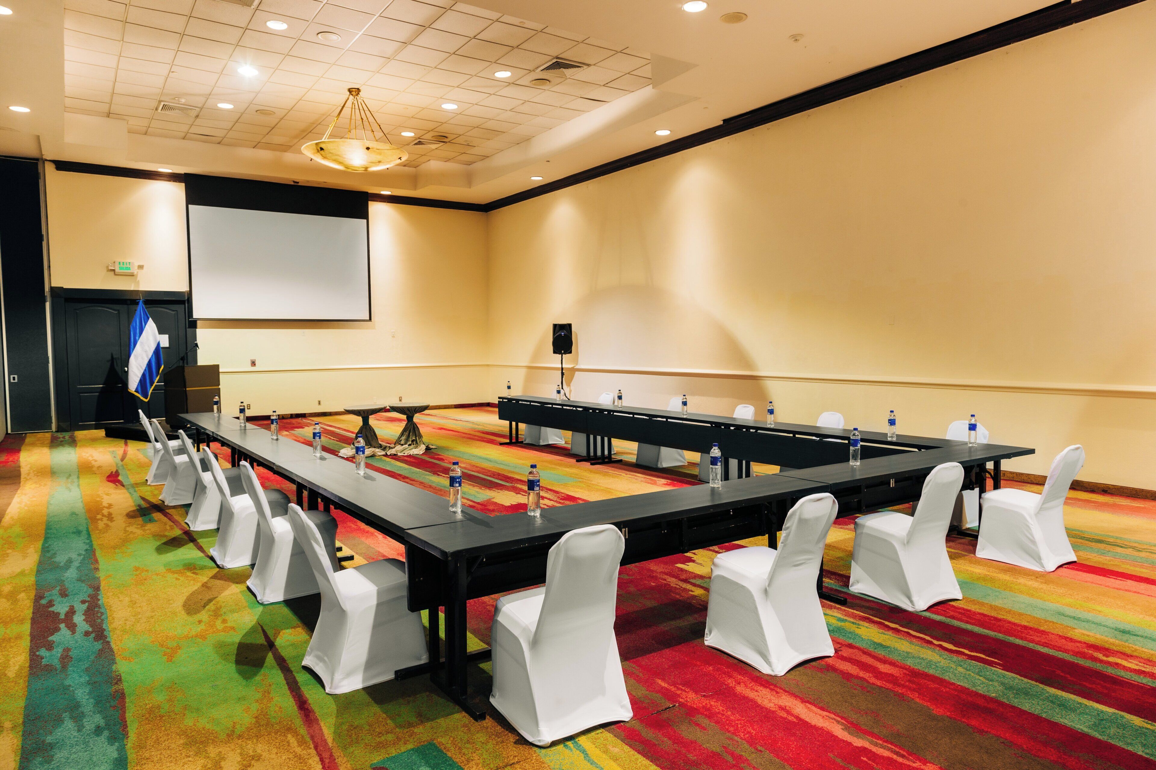 A conference room with a long table, white chairs, and a projector screen.