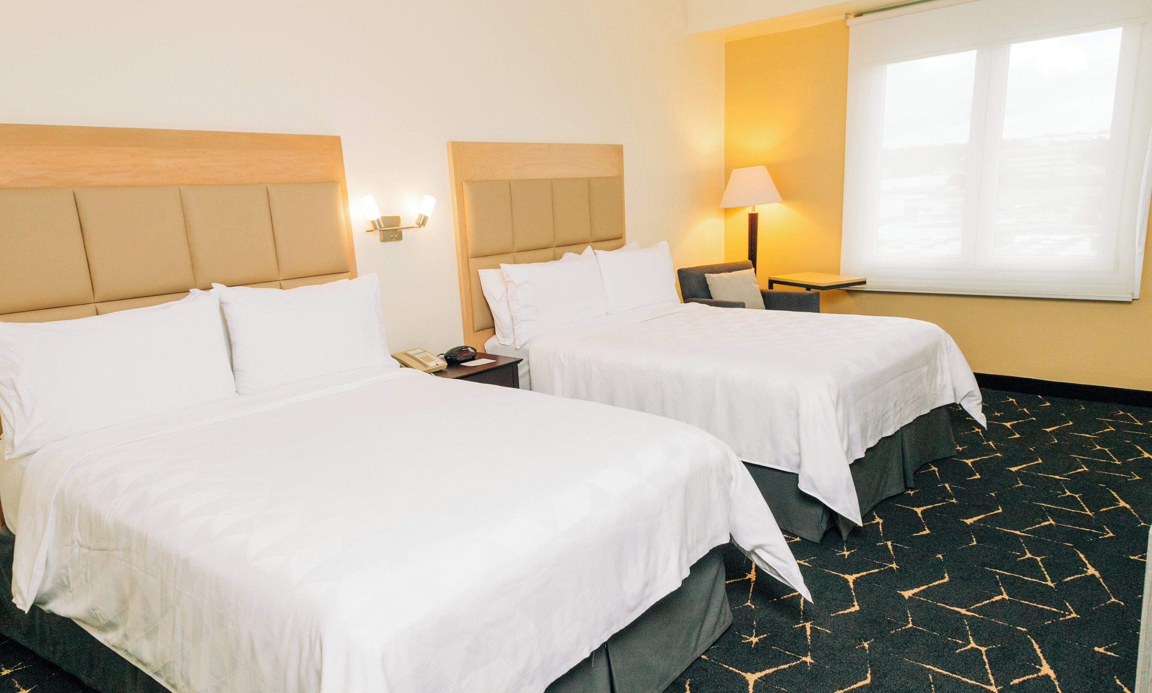 Une chambre d’hôtel avec deux lits, une table de chevet avec un t éléphone, une lampe et une fenêtre avec des rideaux.