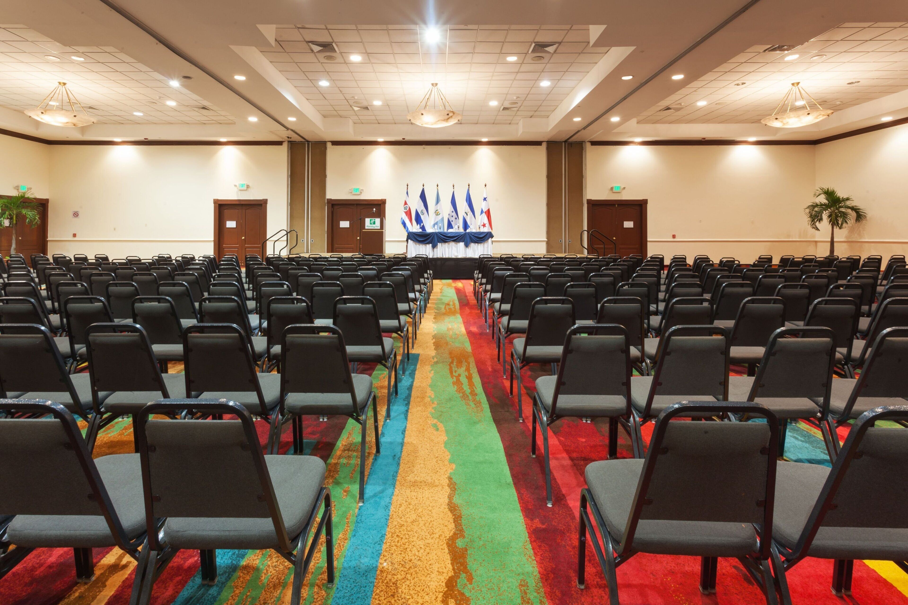 Ein Konferenzraum mit Reihen von Stühlen, die zu einer Bühne mit Flaggen zeigen, einem Teppich mit farbigem Streifen und einer Decke mit eingelassener Beleuchtung.