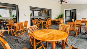 Un espace extérieur spacieux avec des tables et des chaises en bois, un ventilateur de plafond et une entrée avec une porte vitrée.