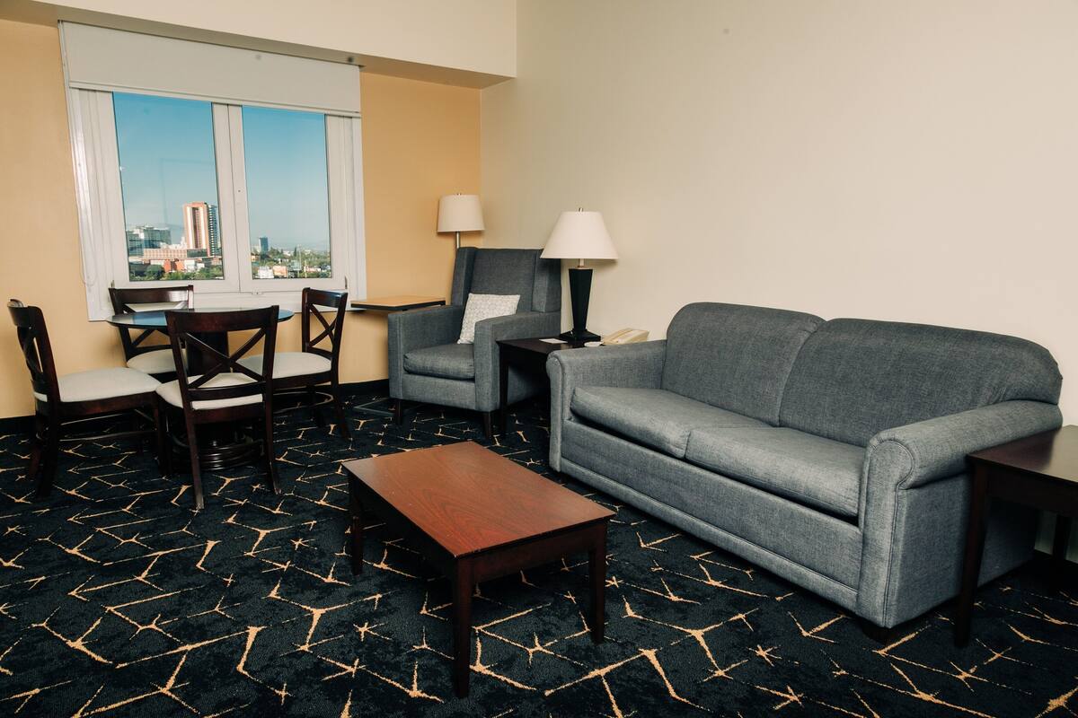 Habitación de hotel con sofá gris, dos sillones, una mesa de centro de madera, una mesa de comedor con sillas y un ventanal que ofrece vistas a la ciudad.