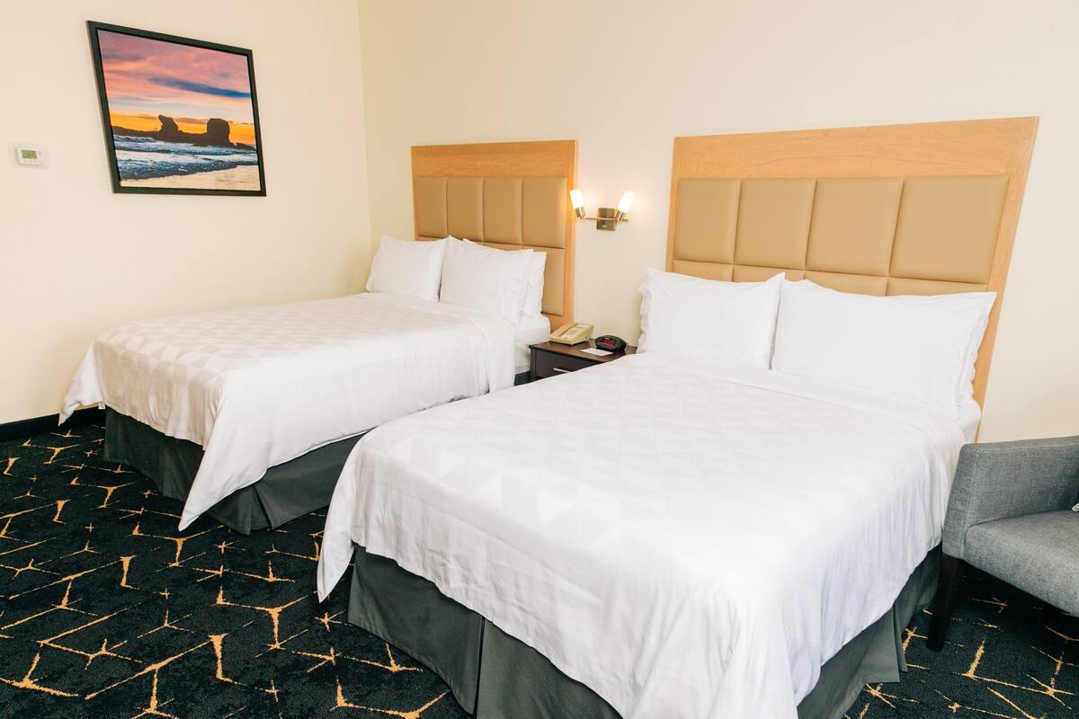 Habitación de hotel con dos camas, un sillón gris, una mesita de noche con un teléfono y un cuadro en la pared.