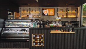 Un café moderne avec un présentoir de pâtisseries, un comptoir où l’on prépare le café et des étagères remplies de boissons.
