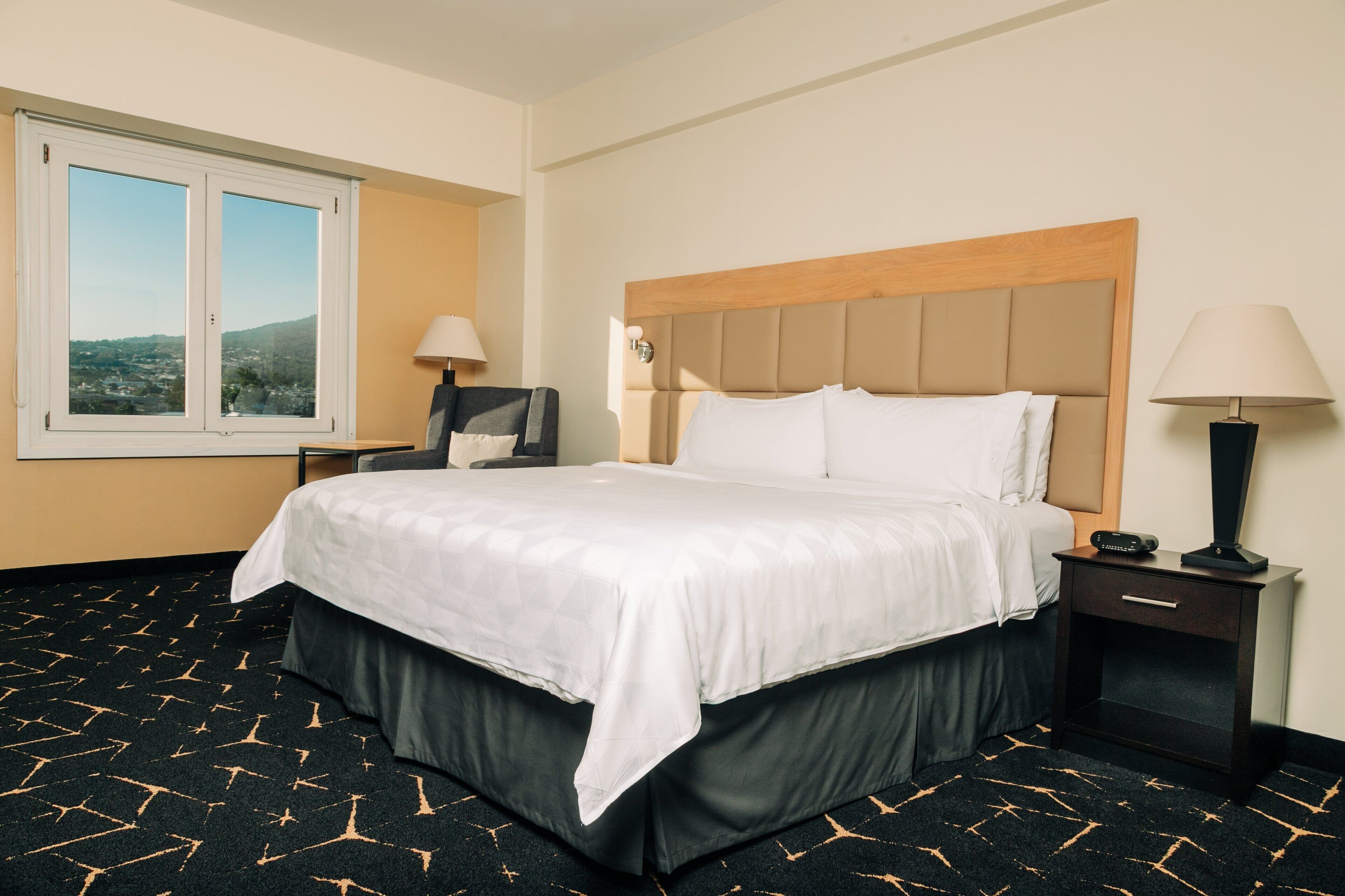 A hotel room with a bed, a nightstand, a lamp, a window, and a view of the outside.