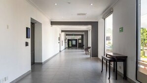 Un long couloir étroit, avec un sol carrelé, un banc et une table sur le côté droit. Des portes se trouvent au fond du couloir, et une fenêtre sur le mur droit.