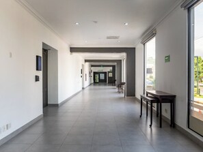 Un pasillo largo y estrecho con suelo de baldosas, un banco y una mesa a la derecha. Hay puertas al final del pasillo y una ventana en la pared derecha.