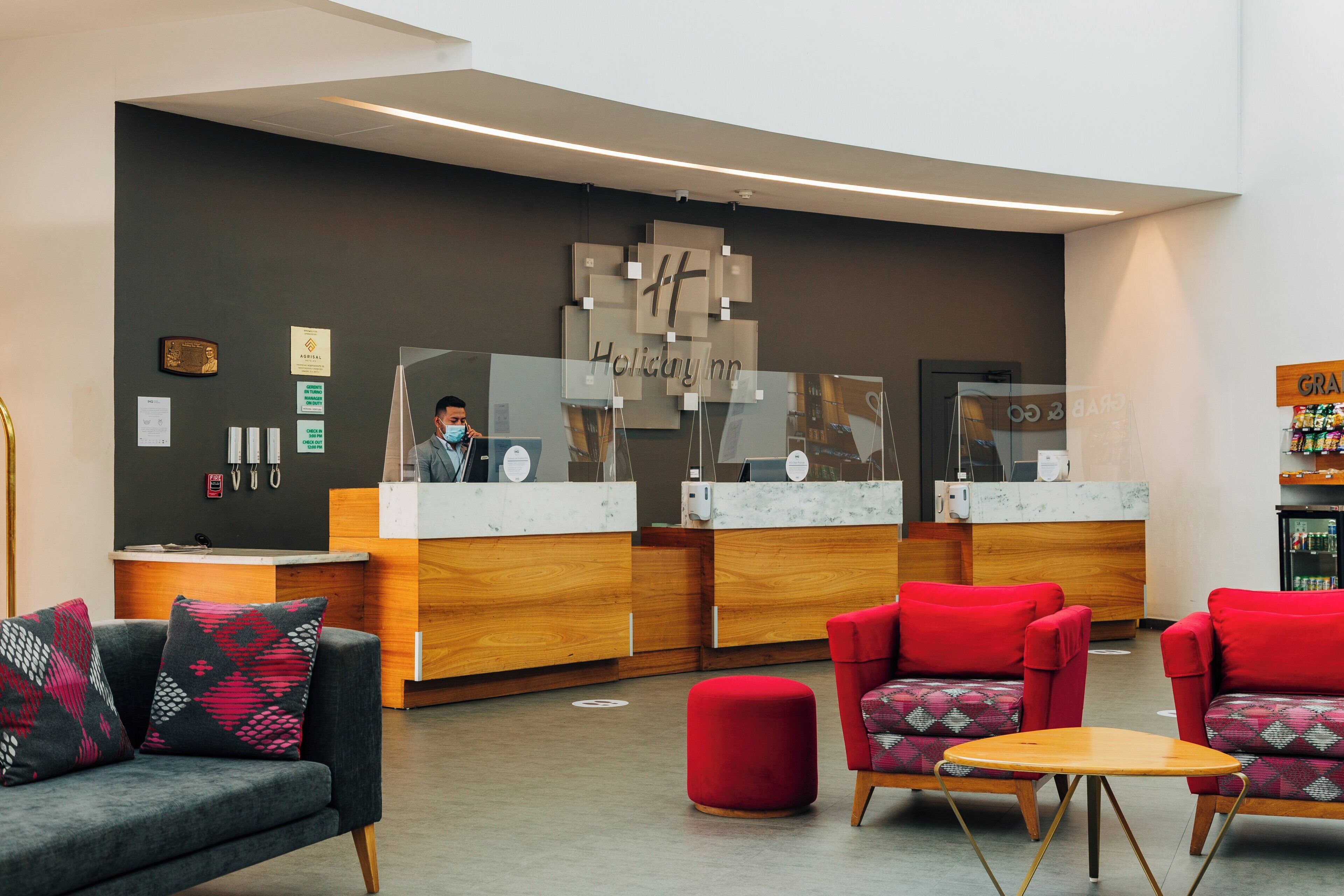Un'area reception di un hotel dal design moderno, con una scrivania della reception, un'area salotto con poltrone imbottite rosse e grigie e una parete con diversi cartelli e un'opera d'arte di grandi dimensioni.
