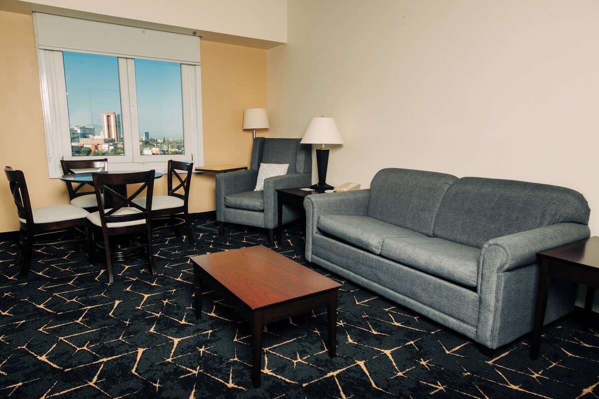 Habitación de hotel con sofá gris, dos sillones, una mesa de centro de madera, una mesa de comedor con sillas y un ventanal que ofrece vistas a la ciudad.