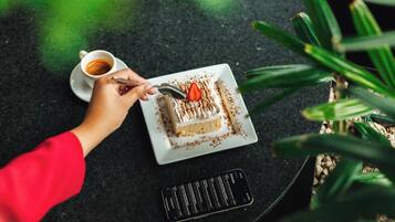 A hand holding a spoon over a slice of cake on a white plate, with a cup of coffee beside it on a dark surface.