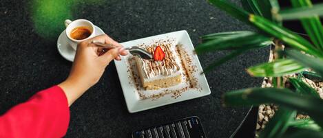 A hand holding a spoon over a slice of cake on a white plate, with a cup of coffee beside it on a dark surface.