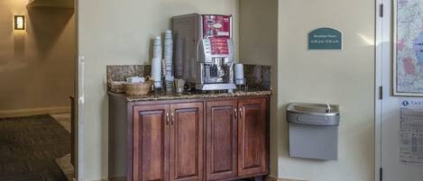 Een ontbijtruimte met een koffieapparaat, een prullenbak en een bord waarop de ontbijttijden staan.